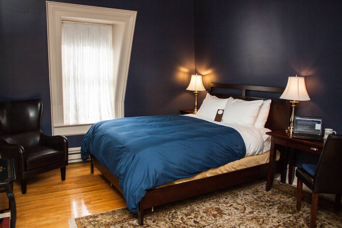Un dormitorio con una cama grande, dos lámparas de noche, una mesita de noche de madera, una computadora portátil, una silla y una ventana con cortinas.