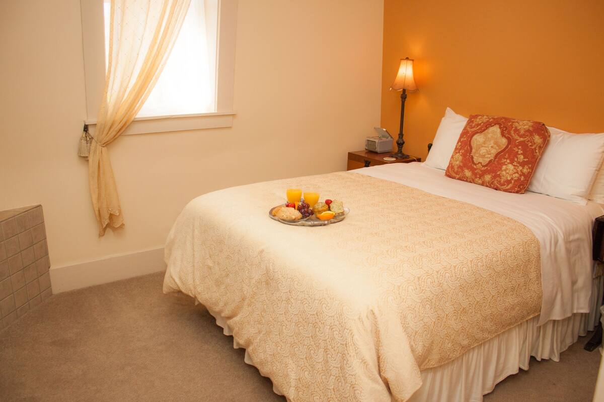 Una cama bien hecha con una bandeja de fruta y una vela, una mesita de noche con una lámpara y una ventana con cortinas.