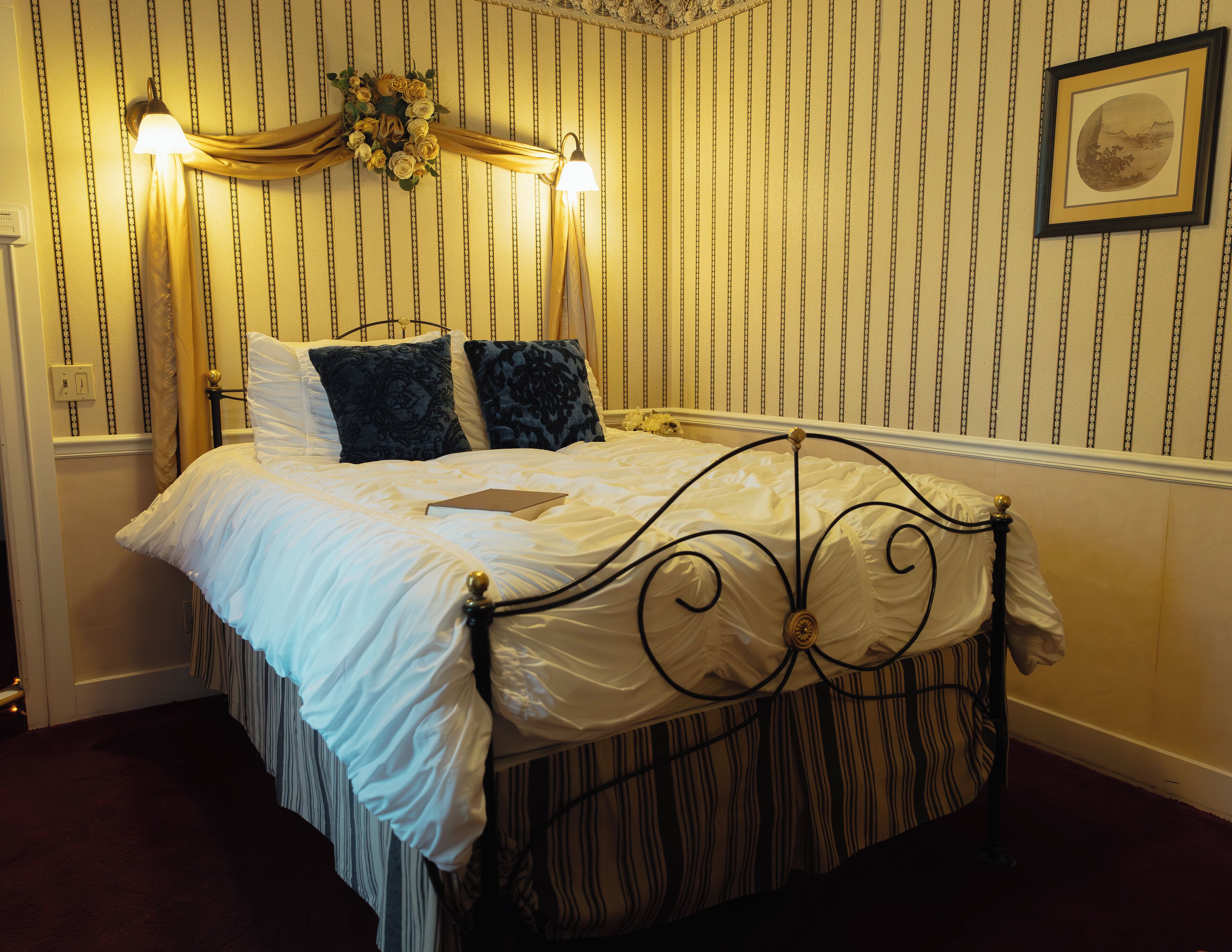 Een netjes opgemaakt bed met een witte deken en twee kussens, een ingelijst beeld aan de muur, en een decoratieve krans boven het bed.
