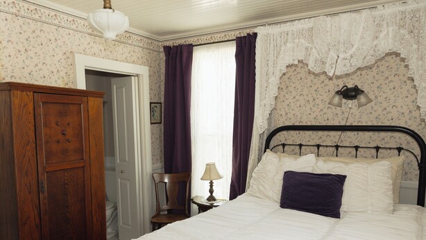 Un dormitorio con un armario de madera, una cama con ropa de cama blanca, una mesita de noche con una lámpara y una cortina de encaje.