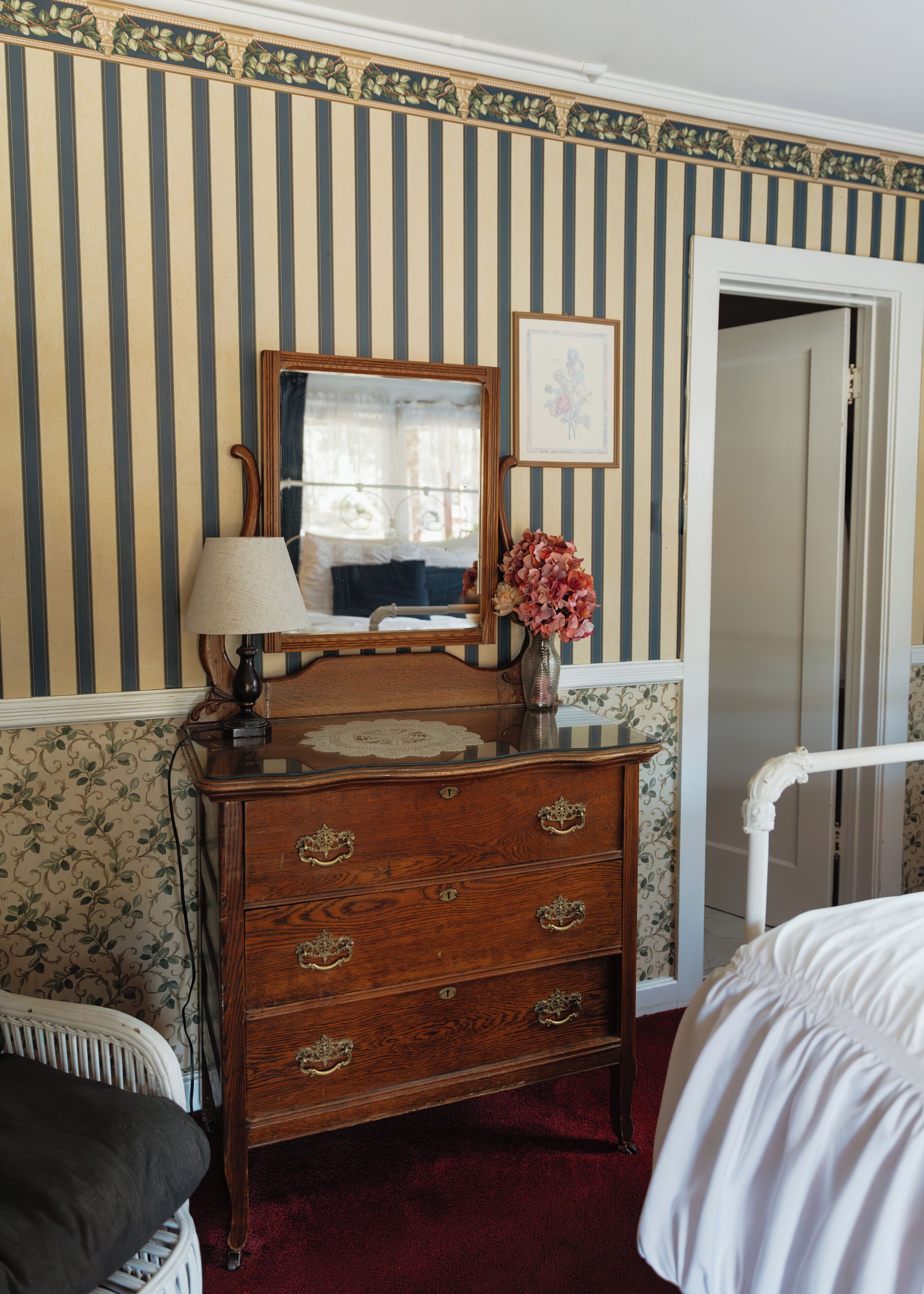 A bedroom with a wooden dresser, a mirror, a bedside table, a lamp, and a vase with flowers.
