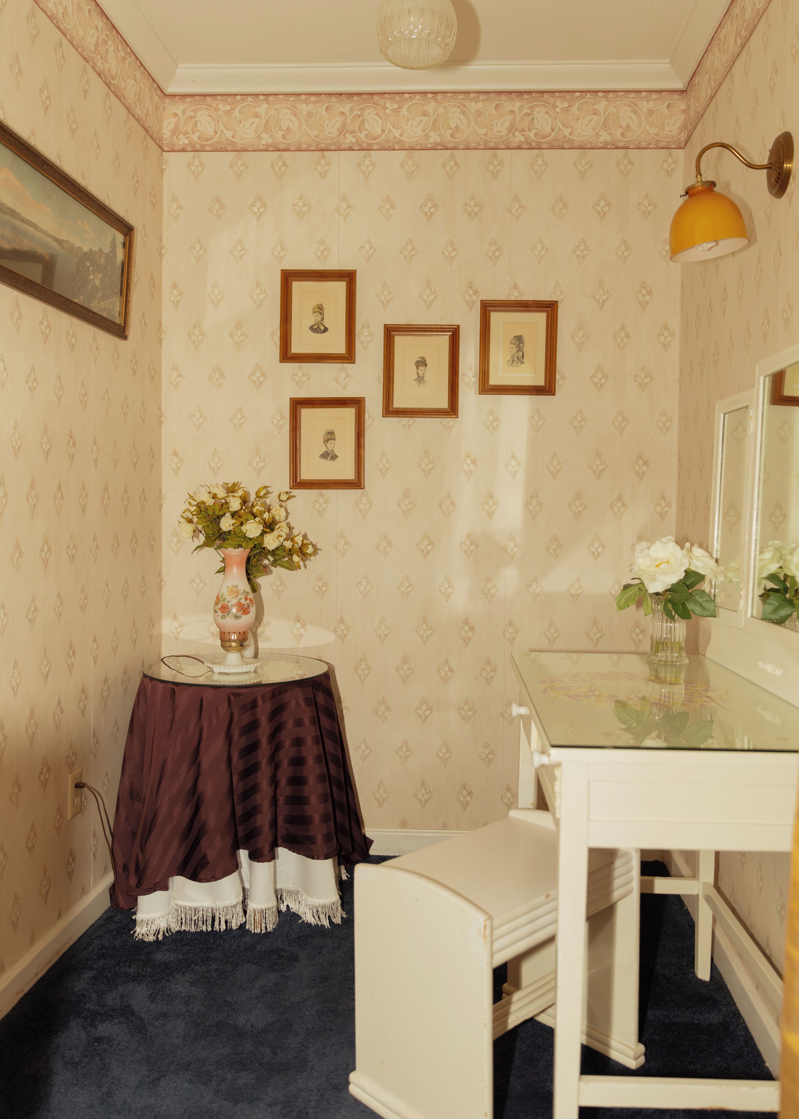 Una habitación con papel tapiz estampado, una pequeña mesa con un jarrón de flores, cuadros en la pared y un espejo sobre la mesa.