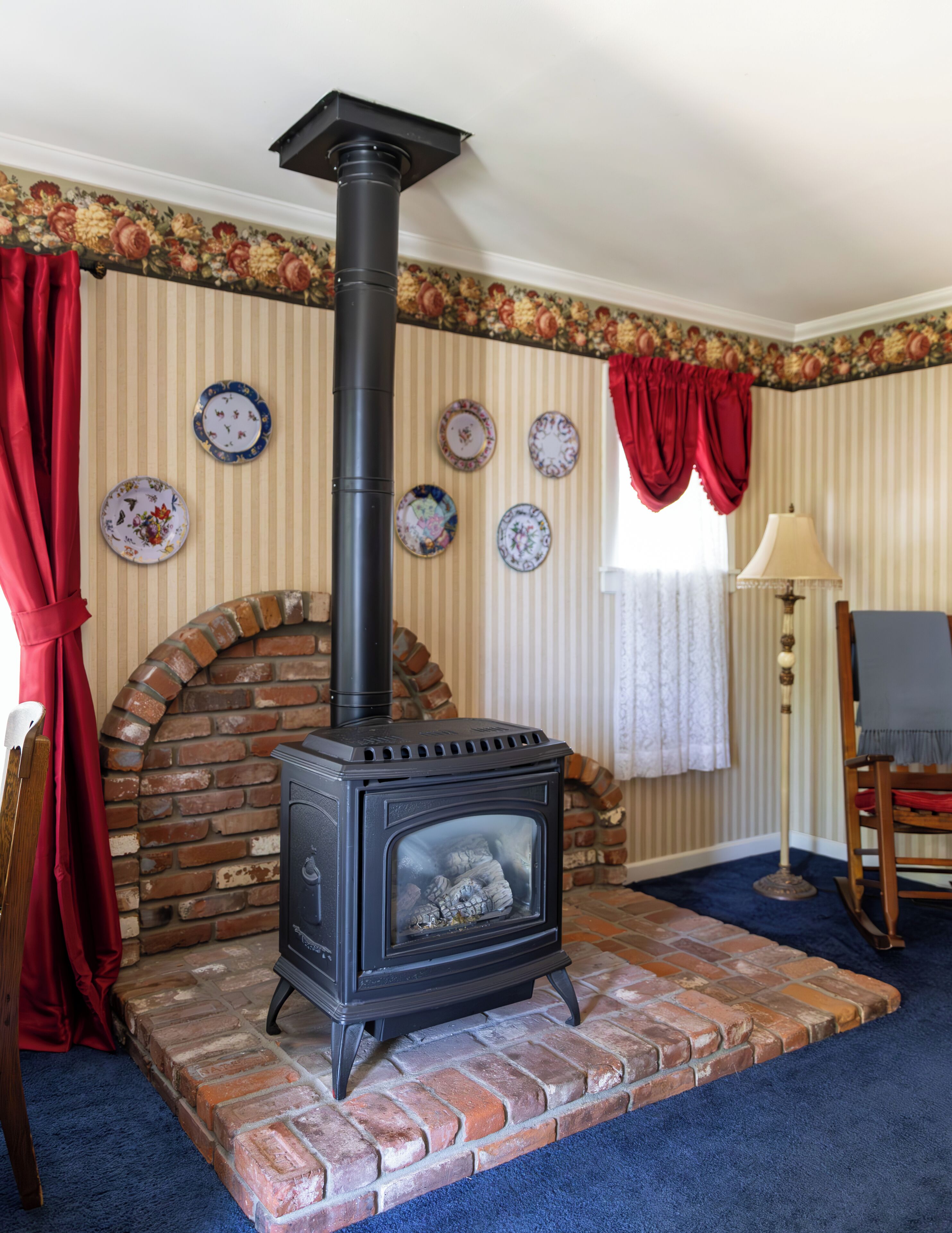 Um quarto com lareira de tijolos, uma cadeira de madeira, um abajur e uma cortina vermelha.