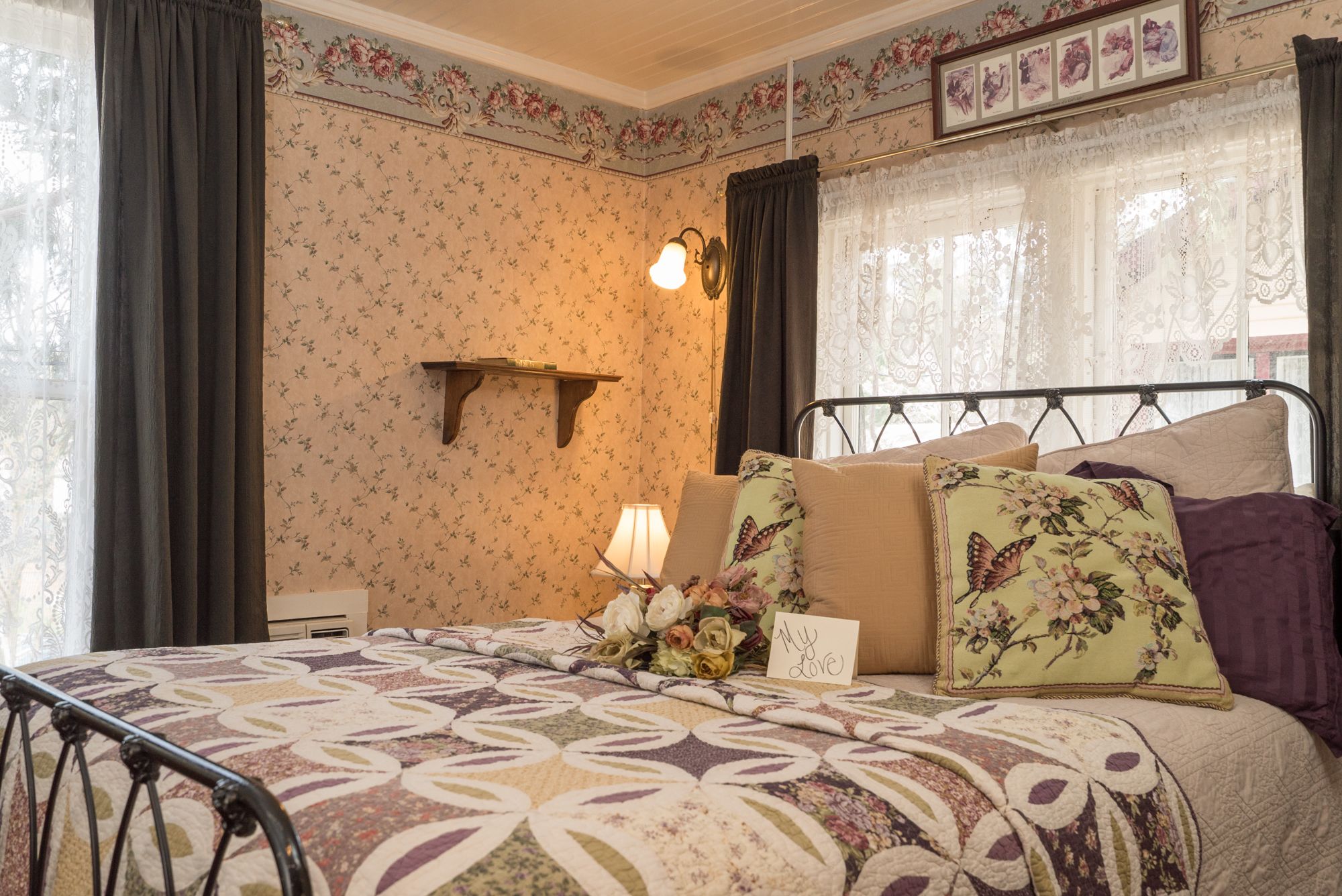Un dormitorio con papel tapiz floral, una cama con ropa de cama estampada, una mesita de noche con una lámpara y un cuadro enmarcado sobre la cama.