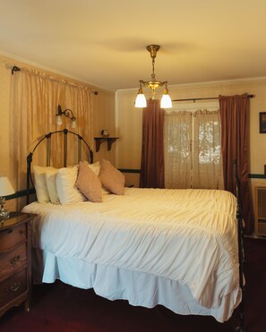 A neatly made bed with a headboard, a bedside table with a lamp, and a window with curtains. - Julian Gold Rush Hotel (Julian)