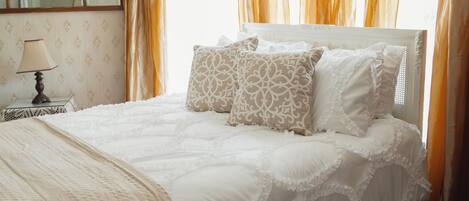 Een net opgemaakt bed met witte lakens en decoratieve kussens, een nachtkastje met een lamp en een spiegel aan de muur.