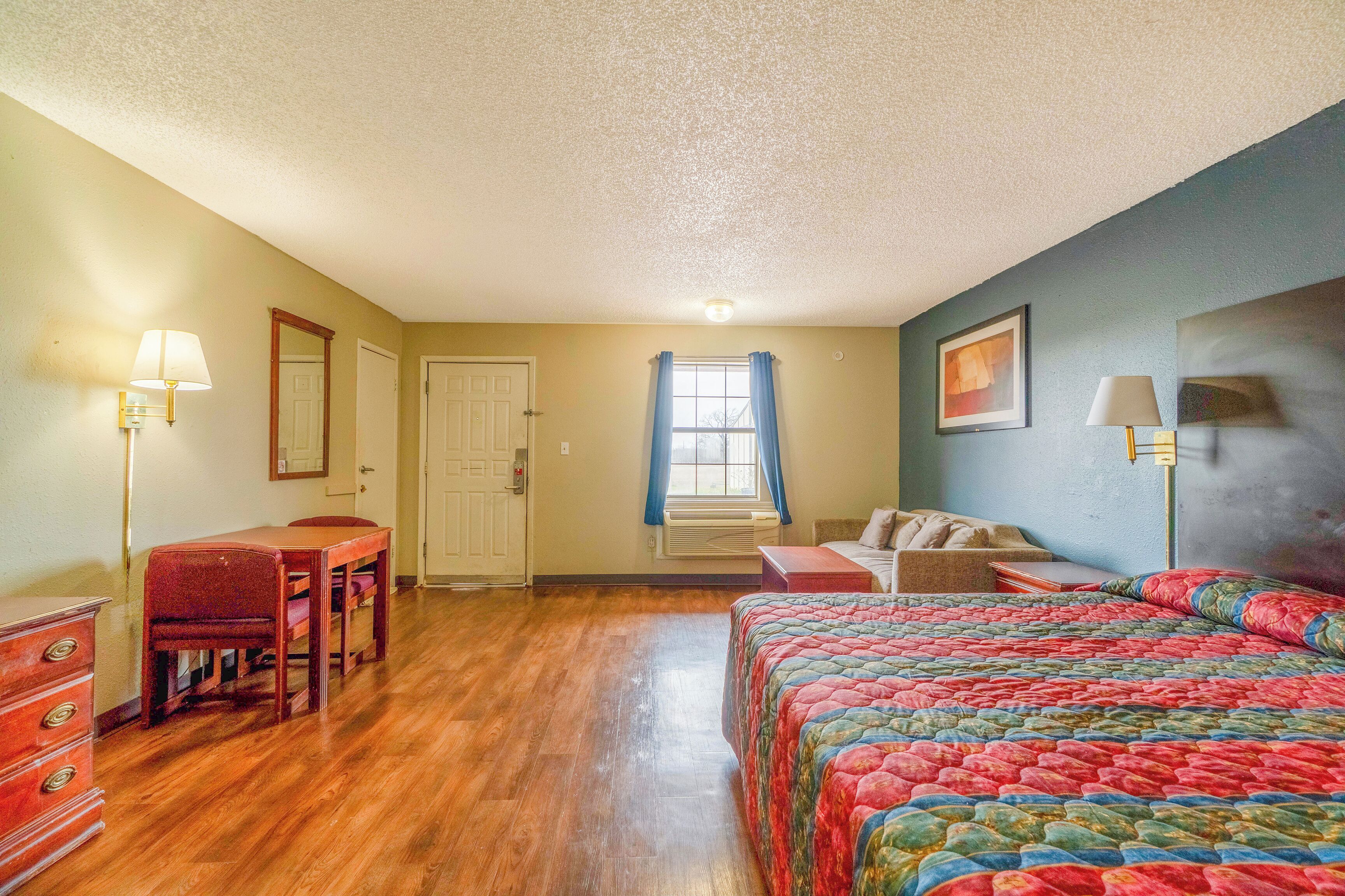 A bedroom with a bed, a desk, a chair, a dresser, a window with curtains, and a painting on the wall.