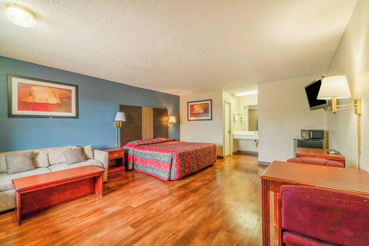 Ein Hotelzimmer mit Holzboden, einer Couch, einem Bett mit bunter Steudecke, einem Schreibtisch, einem Sessel, einem Fernseher und einer Lampe.