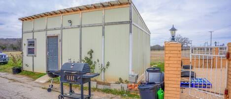 A small, single-story building with a door and windows, a barbecue grill, and a trash bin.