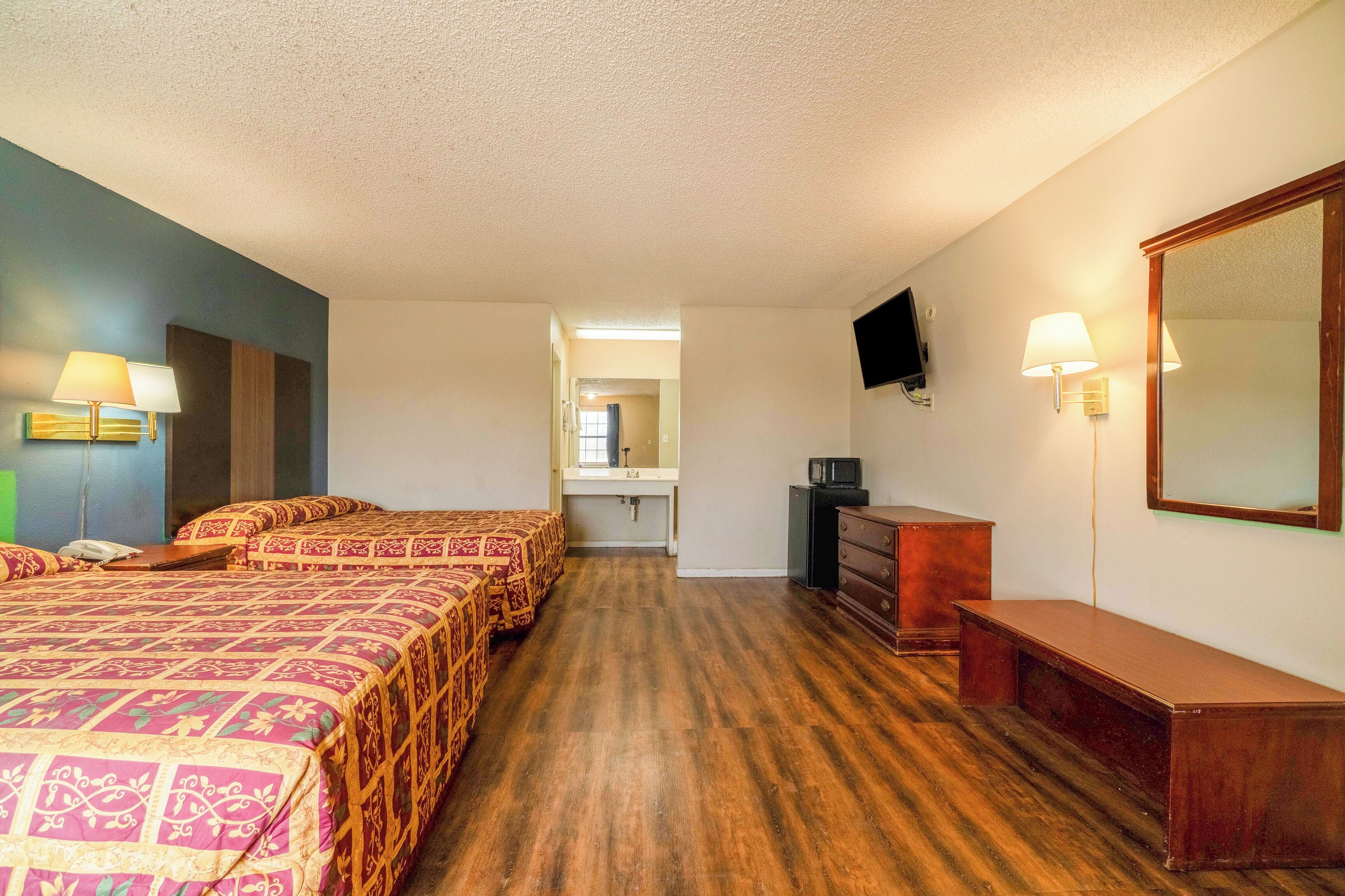 A hotel room with two beds, a desk, a dresser, a TV, and a bathroom in the background.