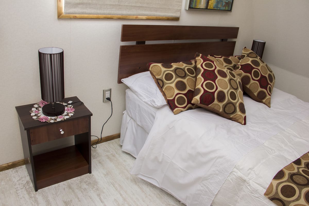 A neatly made bed with patterned pillows, a bedside table with a lamp, and a power outlet on the wall.
