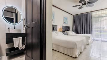Habitación de hotel con puerta de madera oscura, un espejo redondo, un lavamanos blanco y un perchero. La habitación tiene dos camas con ropa blanca, un ventilador de techo y un amplio ventanal con cortinas.