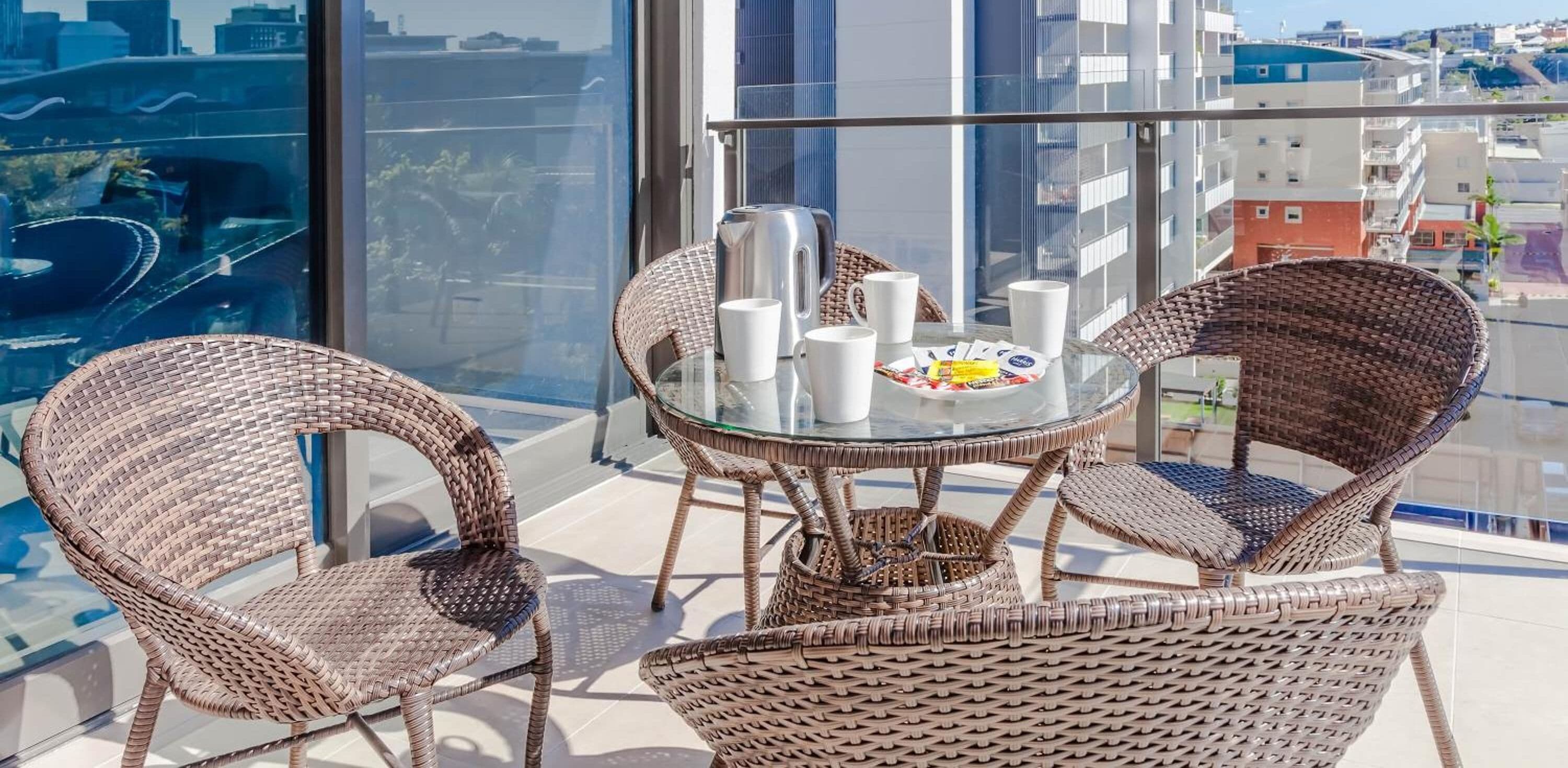 A balcony with wicker chairs and a glass table, overlooking a cityscape.