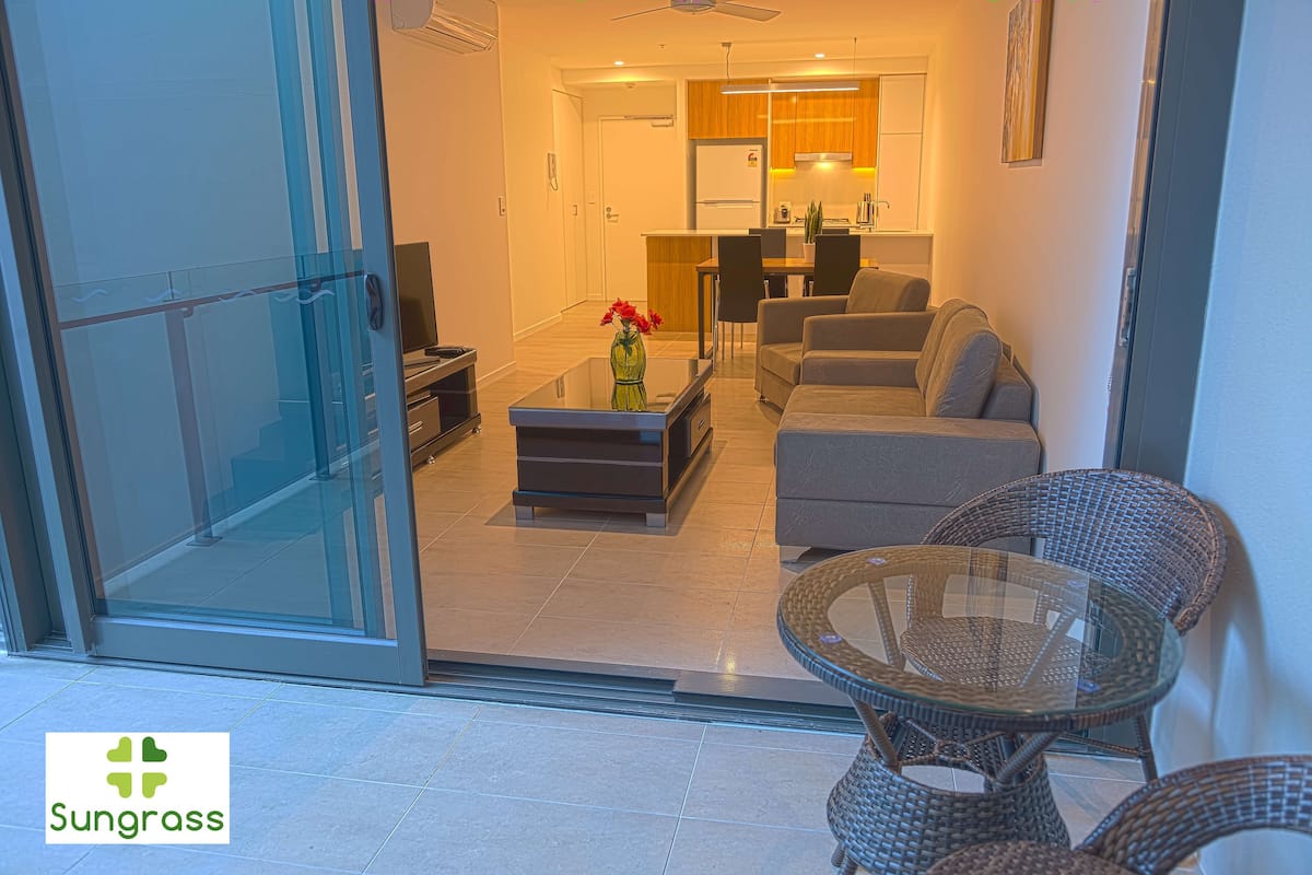 A modern living room with a sofa, a glass coffee table, wicker chairs, and a kitchen in the background.
