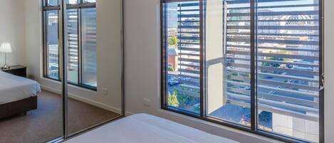 A hotel room with a large bed, a nightstand, a mirror, and a view of a parking garage and buildings through floor-to-ceiling windows.