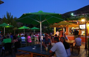 An outdoor dining area with tables and chairs under umbrellas, a bar area with people, and a mountainous backdrop.