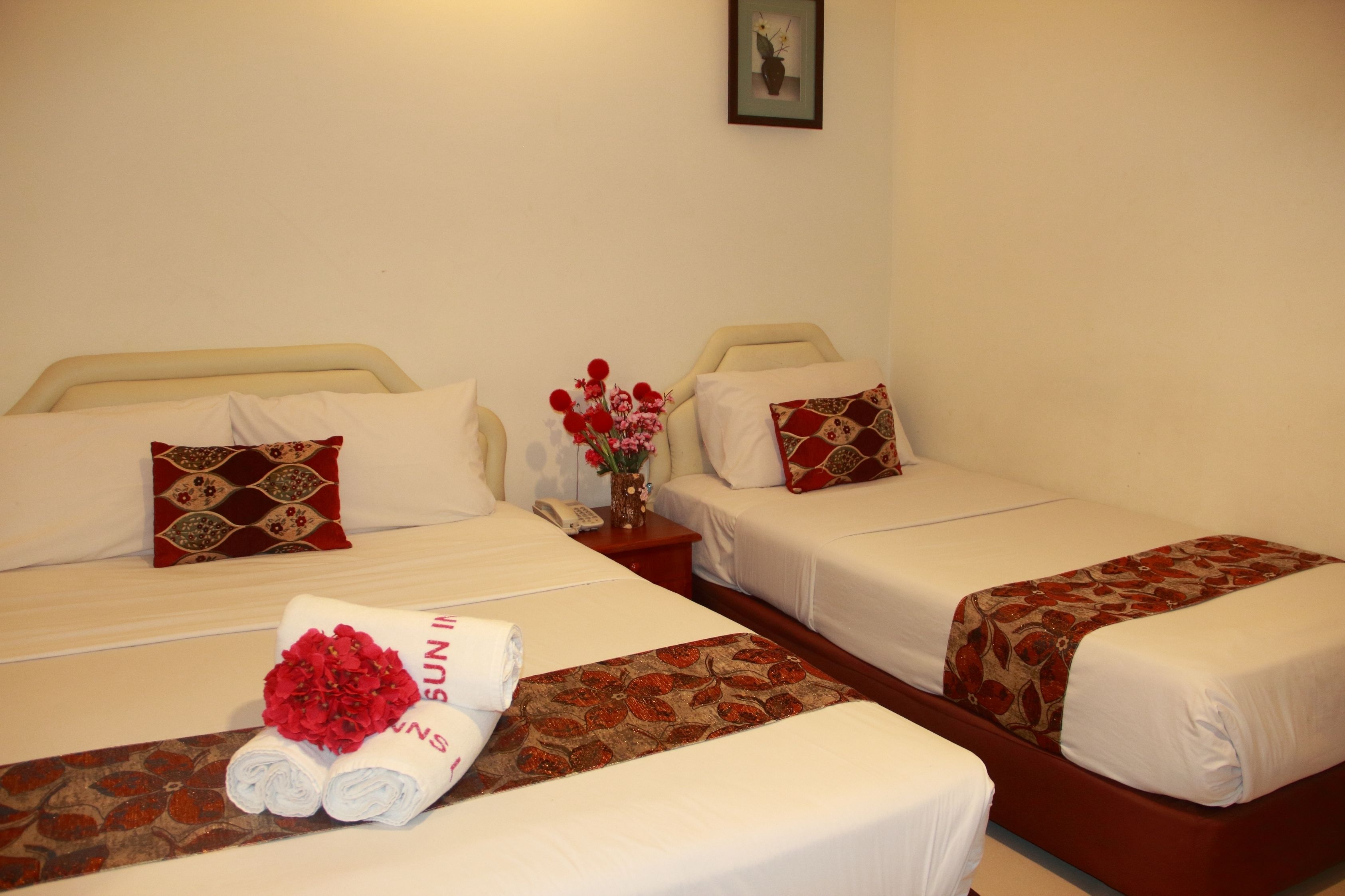 A hotel room with two single beds, each with a patterned bedspread and matching pillows. There is a small table with a vase of flowers between the beds.