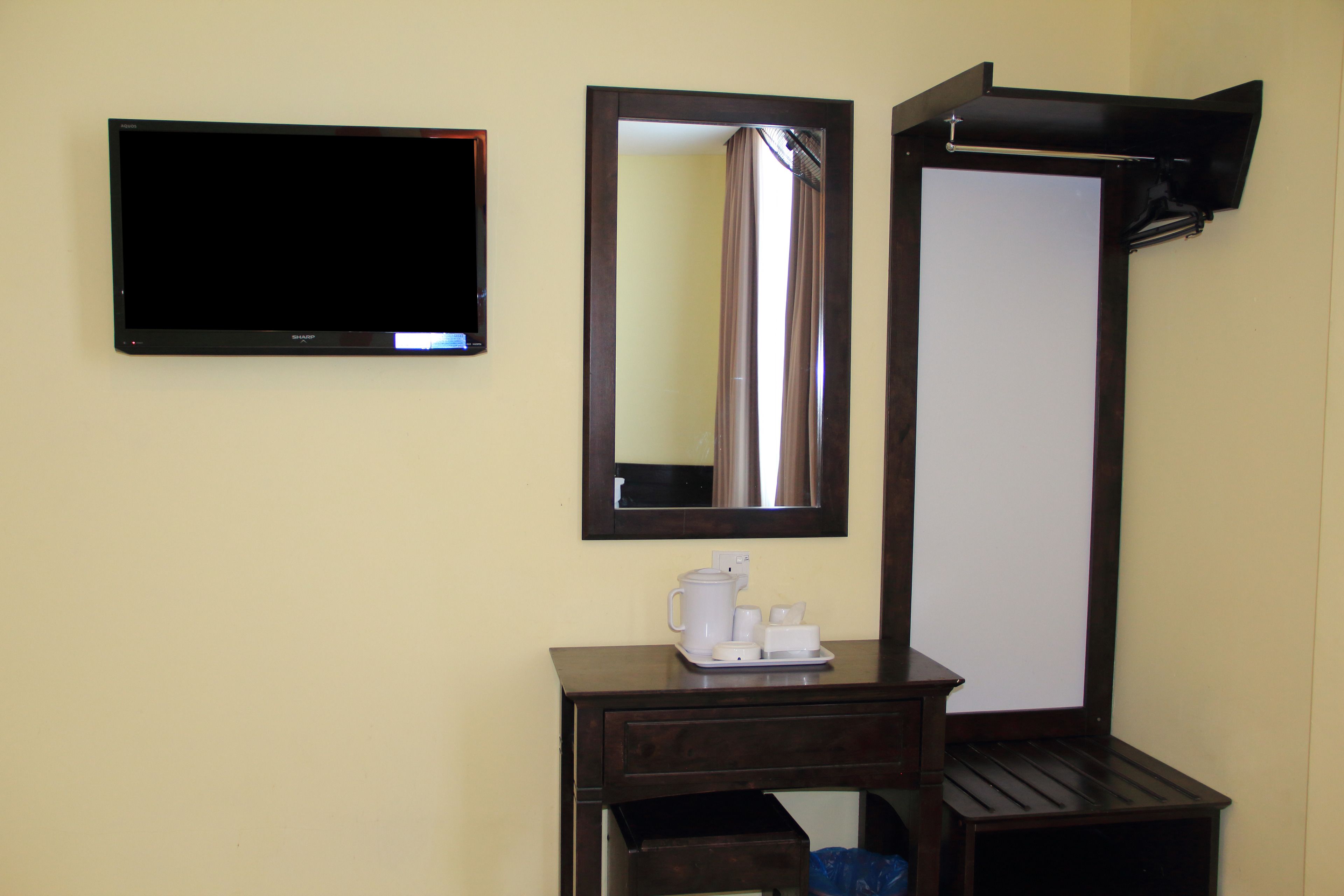 A hotel room with a flat-screen TV, a large mirror, a small desk with a coffee maker, and a trash bin.