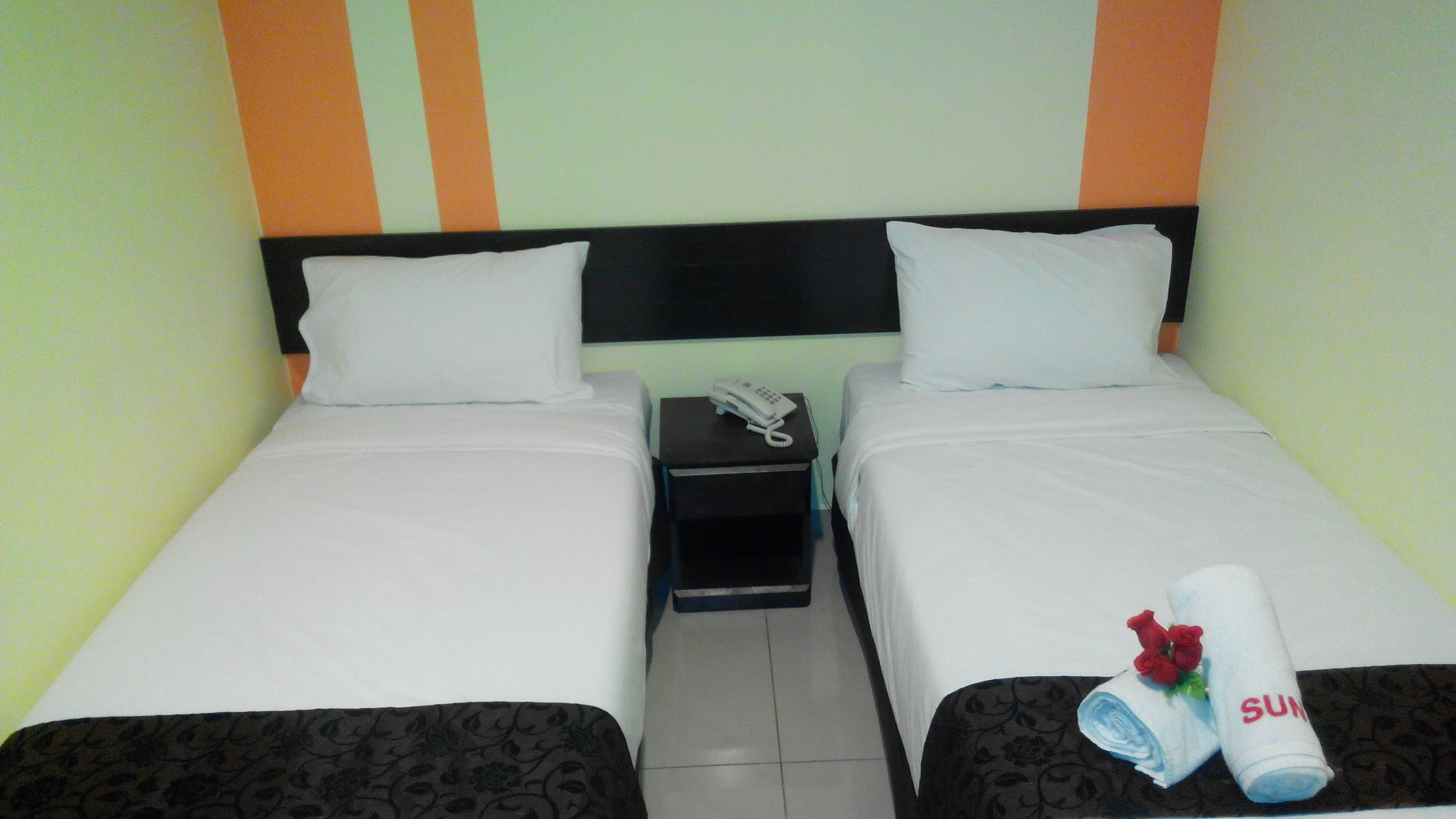 A twin room with two beds, a bedside table with a phone, and a towel with a rose and the word "SUN" on it.