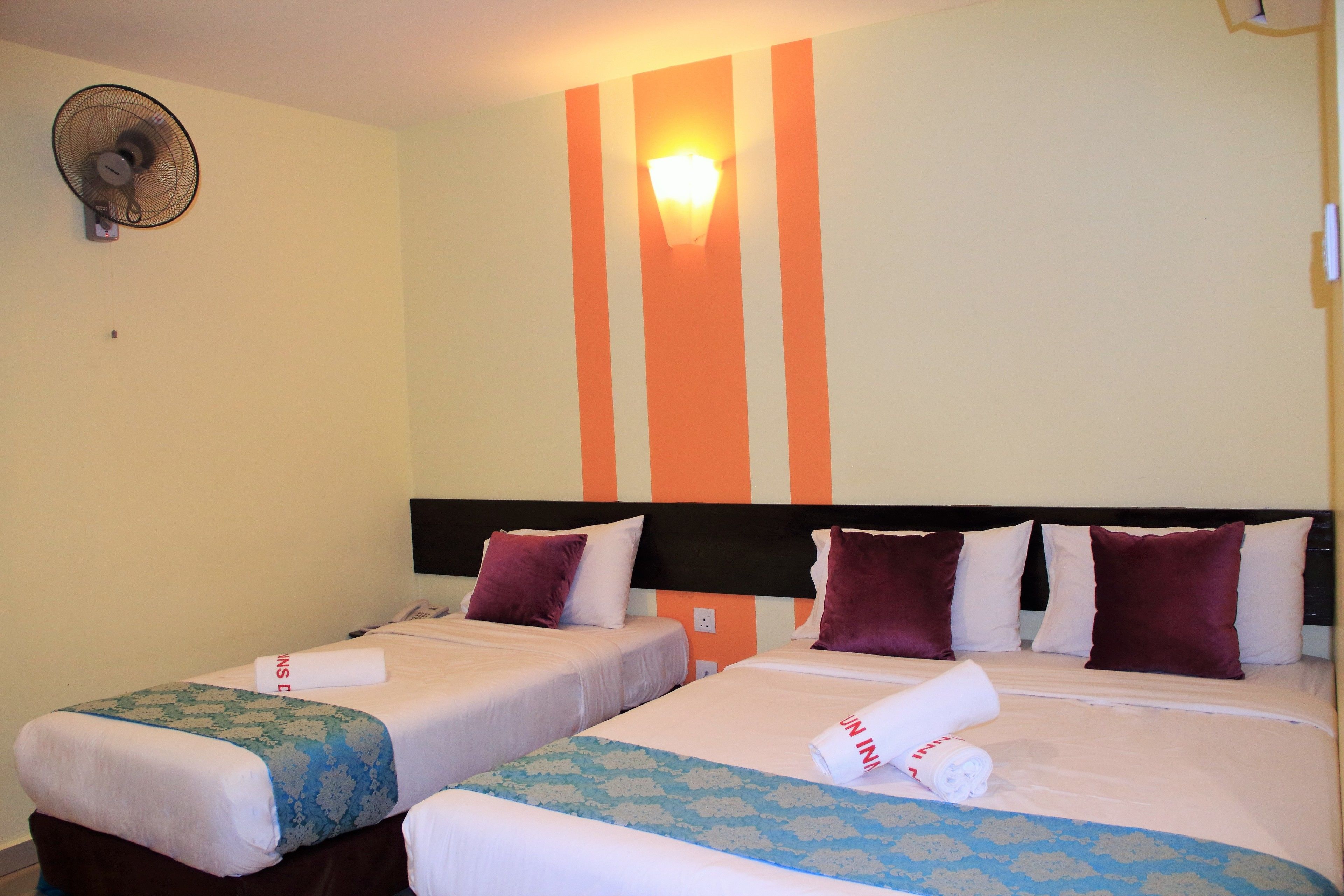 Une chambre d’hôtel avec deux lits simples, un ventilateur fixé au mur et un luminaire.