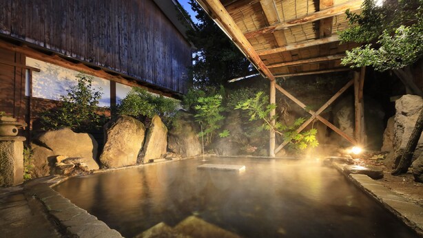 Un onsen japonés tradicional con estructura de madera, escalones de piedra y un baño de piedra rodeado de rocas y vegetación.