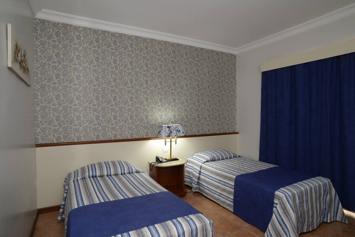 Una camera d'albergo con due letti singoli, un comodino con una lampada e carta da parati decorata.