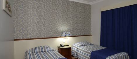Habitación de hotel con dos camas individuales, una mesita de noche con lámpara y papel tapiz estampado.