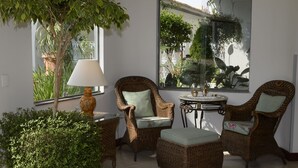 Un coin salon accueillant, avec des fauteuils en osier et une table, entouré de plantes en pot et d’une grande fenêtre donnant sur un paysage verdoyant.