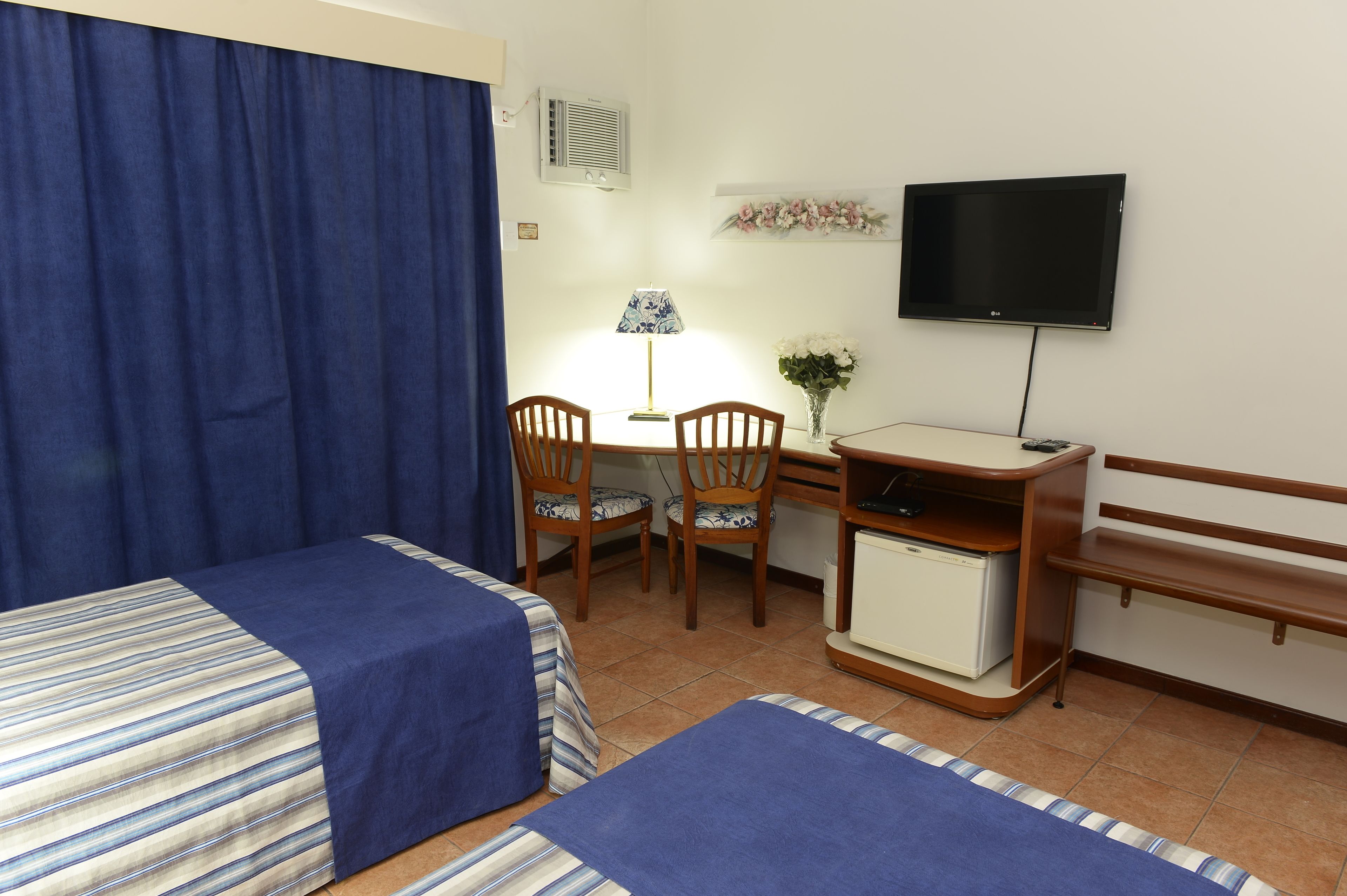 Habitación de hotel con dos camas, un escritorio con lámpara, televisión y refrigerador pequeño.