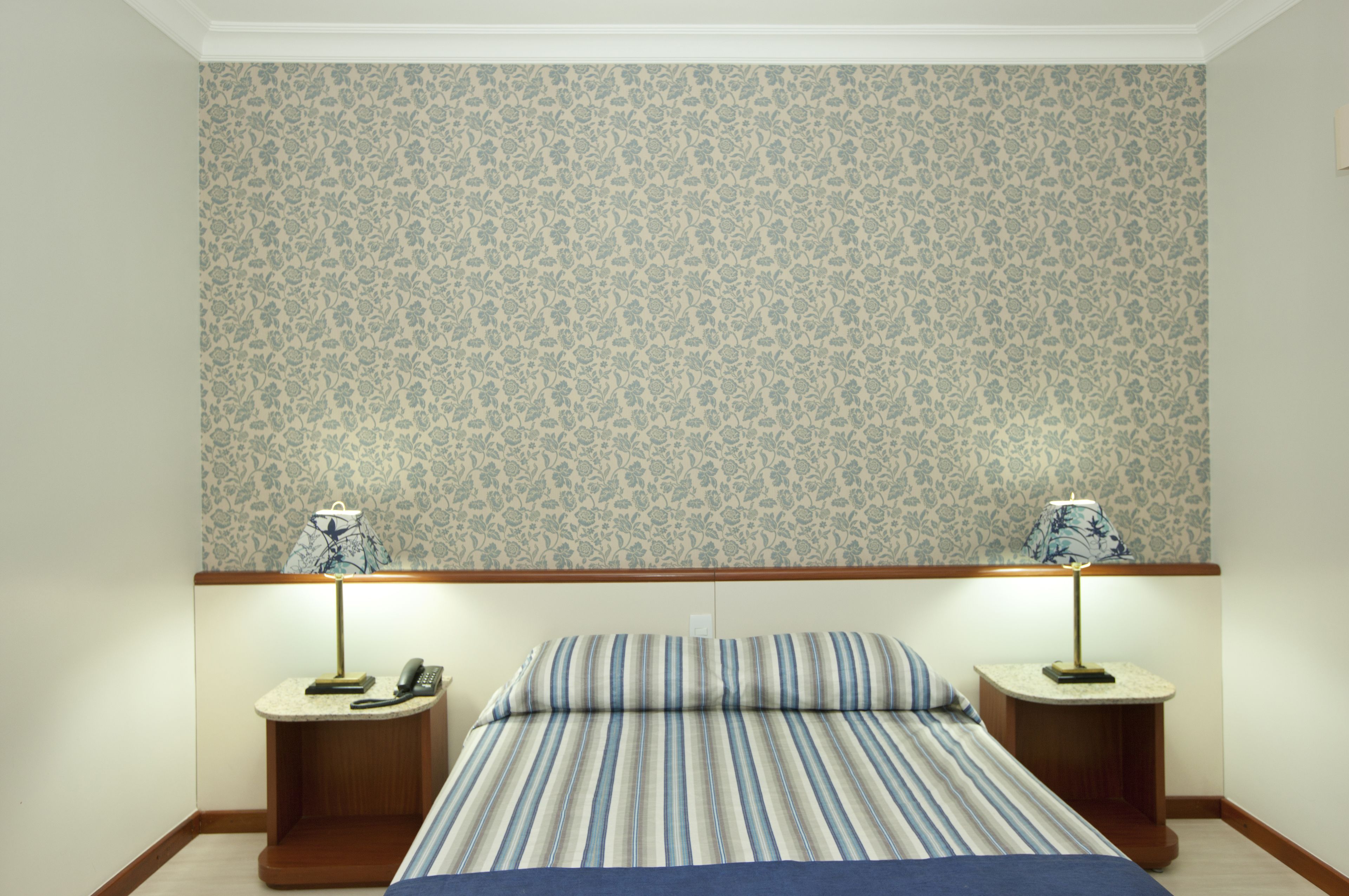 Een slaapkamer met behang met bloemmotief, een streepjesbedovertrek, twee nachtkastjes met lampen en een telefoon op één van de tafels.