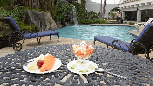 Une table placée au bord de la piscine, sur laquelle se trouvent un saladier de fruits et des assiettes de fruits, entourée de chaises longues et donnant sur une piscine avec une cascade en arrière-plan.