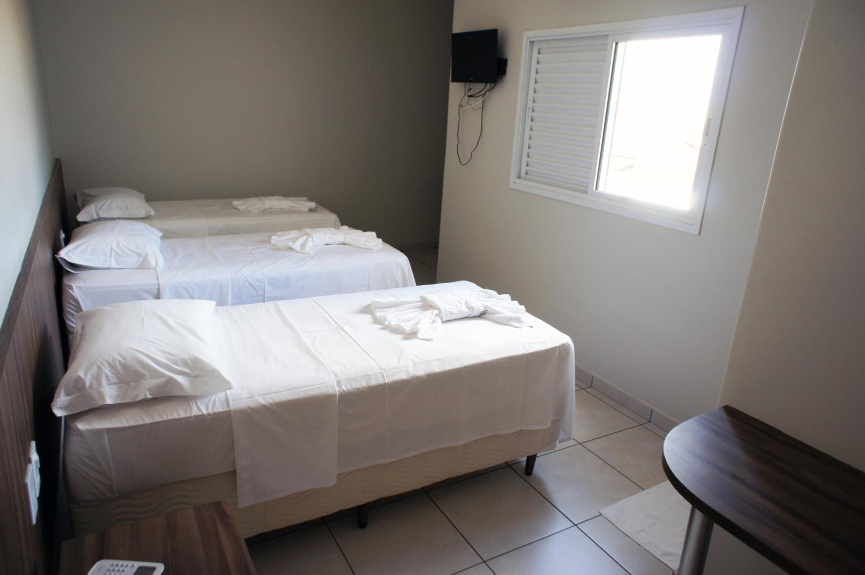 Quarto com duas camas de solteiro, uma mesinha, televisão na parede e janela com persianas.
