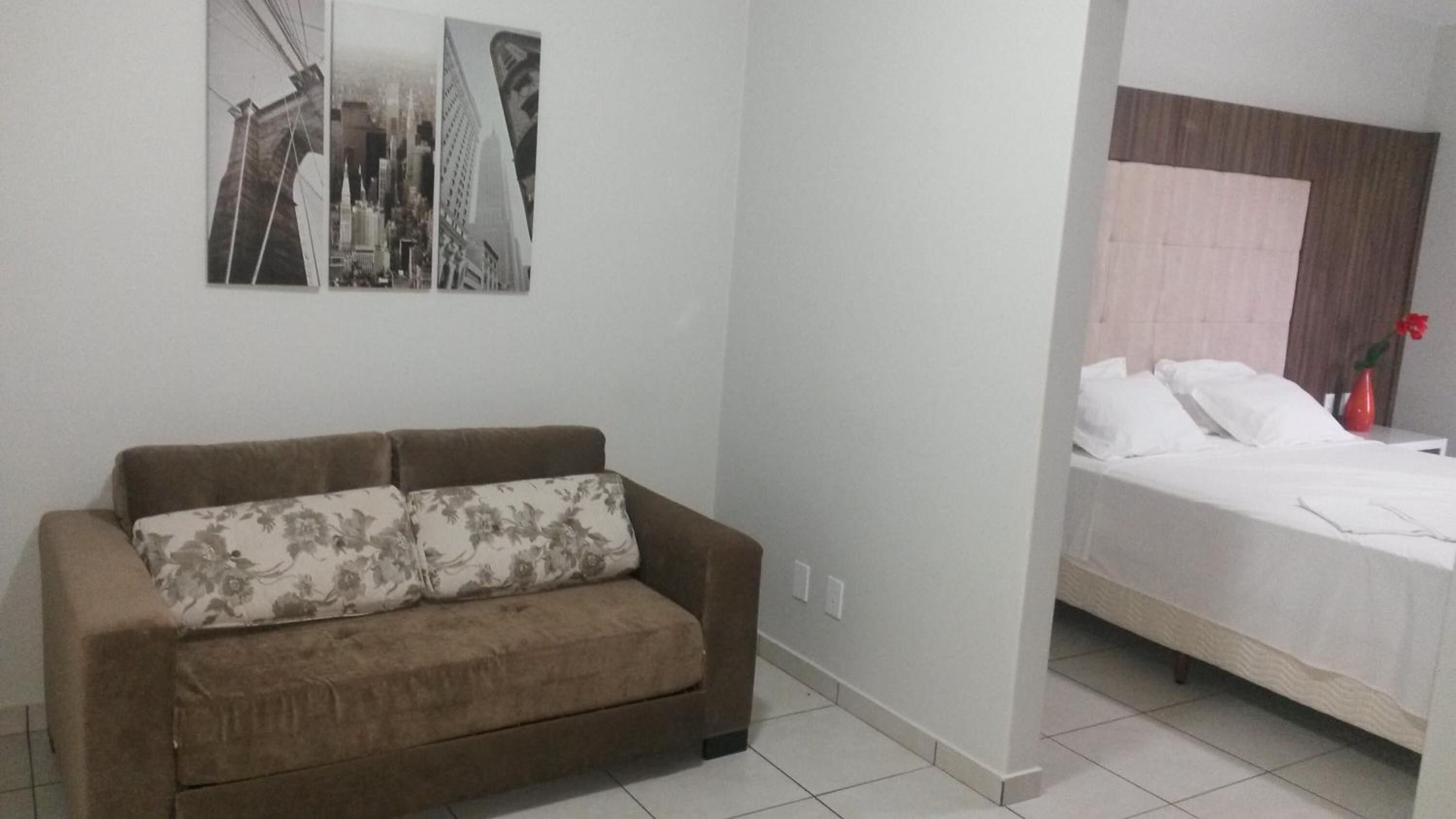 Una habitación con un sofá marrón, una cama con ropa de cama blanca y una obra de arte colgada en la pared que muestra edificios de la ciudad.