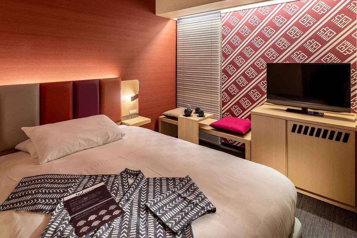Ein Hotelzimmer mit Bett, Fernseher, Schreibtisch und Fenster mit Jalousien.
