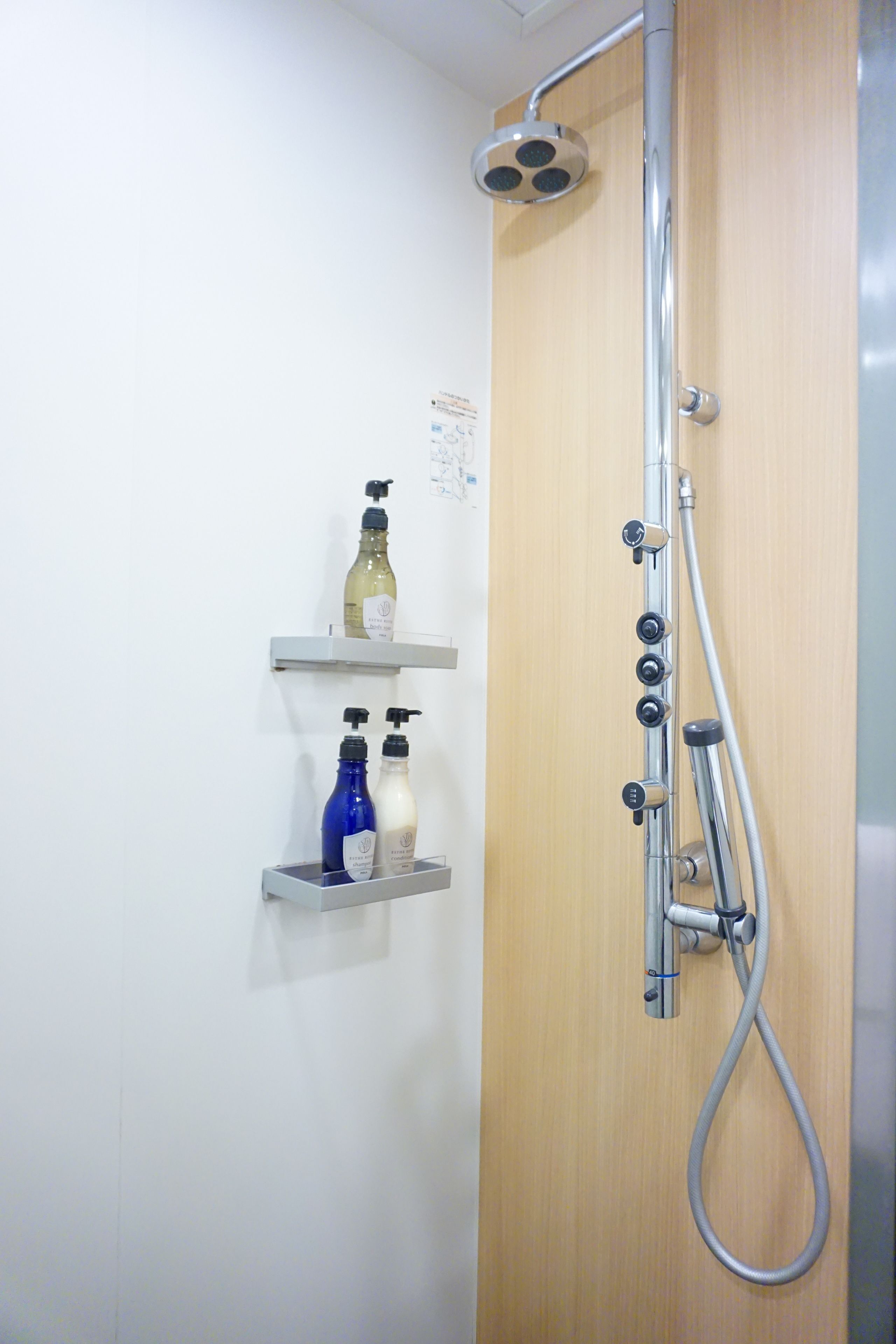 Une douche équipée d’un pommeau de douche à main et d’un flexible. On y trouve deux étagères avec des bouteilles de shampoing et d’après-shampoing.