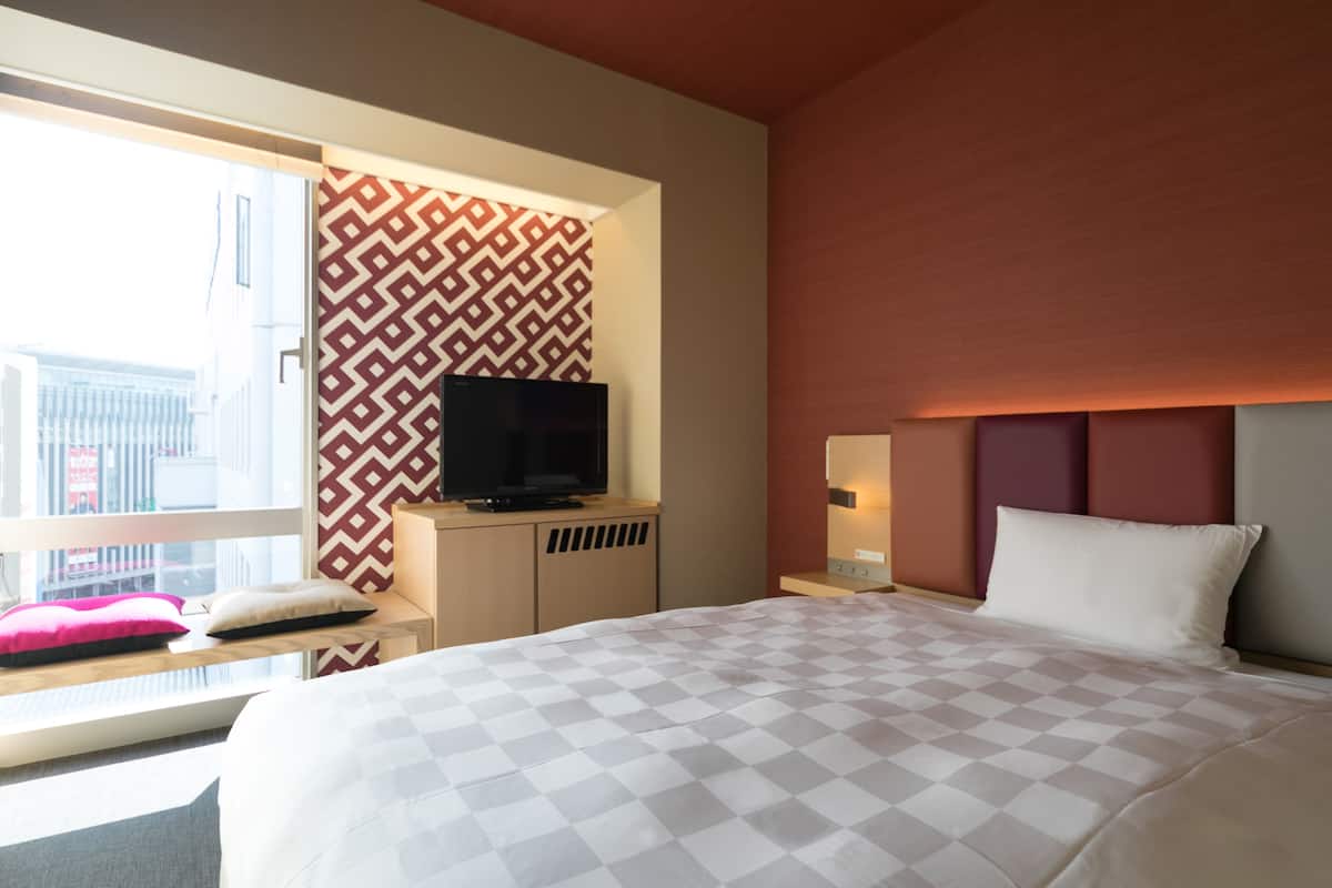 Une chambre d’hôtel avec un grand lit, un téléviseur posé sur un meuble en bois, une fenêtre donnant sur des immeubles et un mur orné d’un motif.