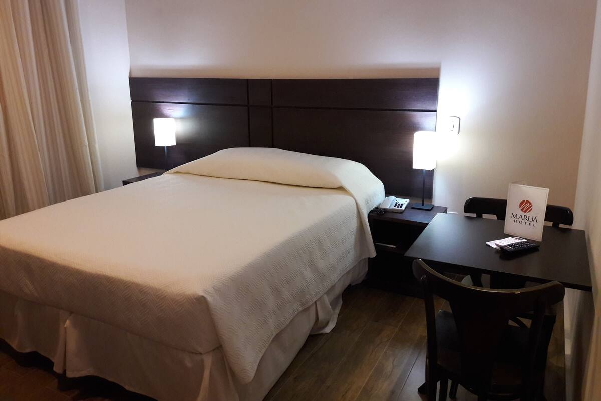 Ein Hotelzimmer mit Bett, Nachttisch, Lampe, Stuhl und einem Schild mit dem Hotelnamen.