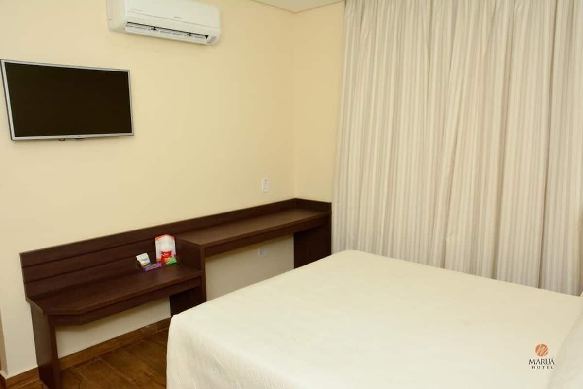 Ein Hotelzimmer mit Bett, Wandfernseher, Schreibtisch mit Stuhl und einem fensterbepflanzten Fenster.