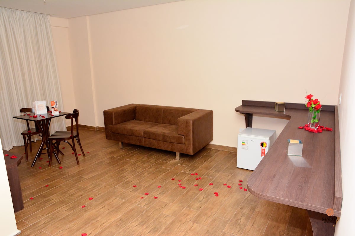 Ein Zimmer mit einem braunen Sofa, einem Esstisch mit Stühlen, einem kleinen Kühlschrank und einer Holzbank.