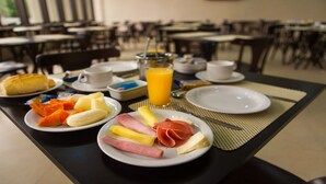 A breakfast setup with plates of food, a glass of orange juice, and a cup of coffee on a table in a dining area with chairs and tables. - Hotel Maruá (São José dos Campos)