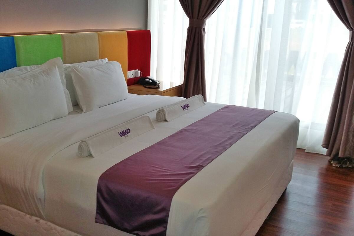Une chambre d’hôtel avec un grand lit, une tête de lit ornée de panneaux colorés, une table de chevet avec un téléphone et une fenêtre avec des rideaux.