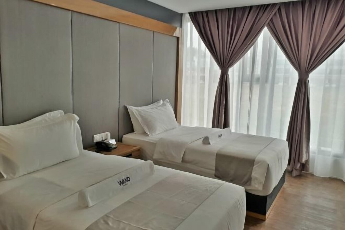 A hotel room with two beds, a wooden headboard, a nightstand with a phone, and sheer curtains.
