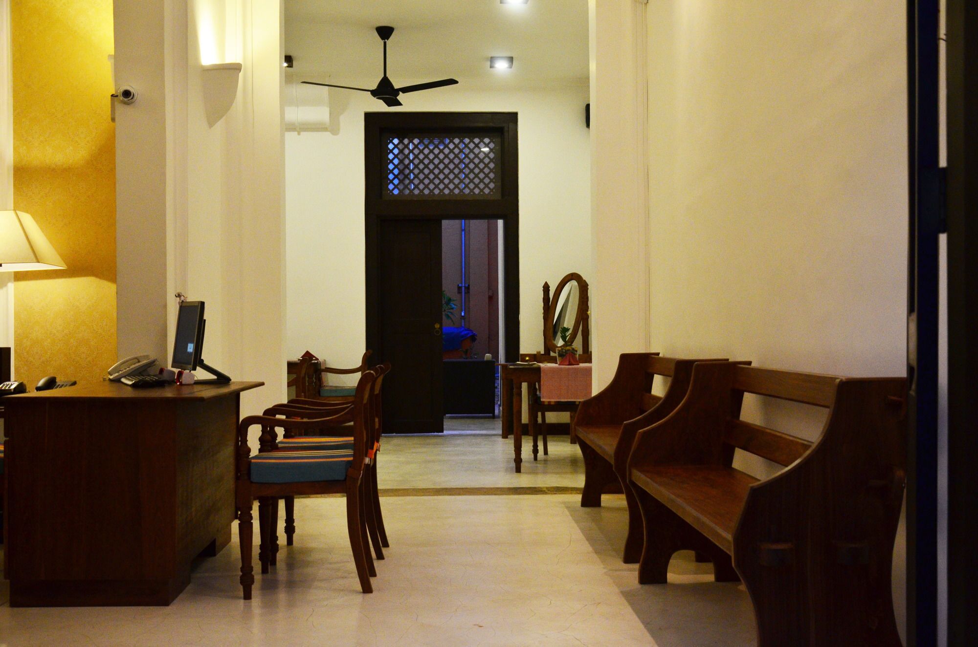A hotel lobby with a reception desk, several wooden chairs, a bench, a ceiling fan, and a door with a decorative screen.