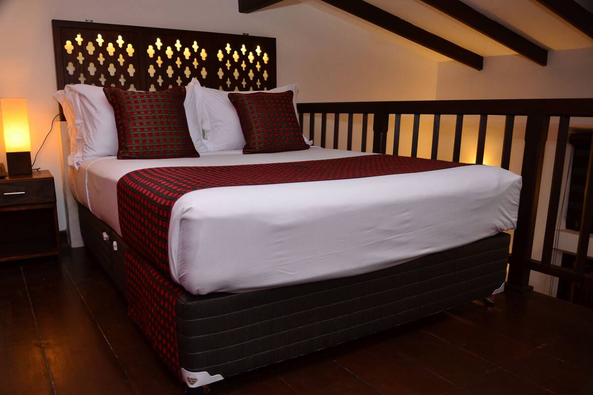 A neatly made bed with a patterned bedspread and pillows, a bedside lamp, and a dark wooden headboard with a decorative light panel.