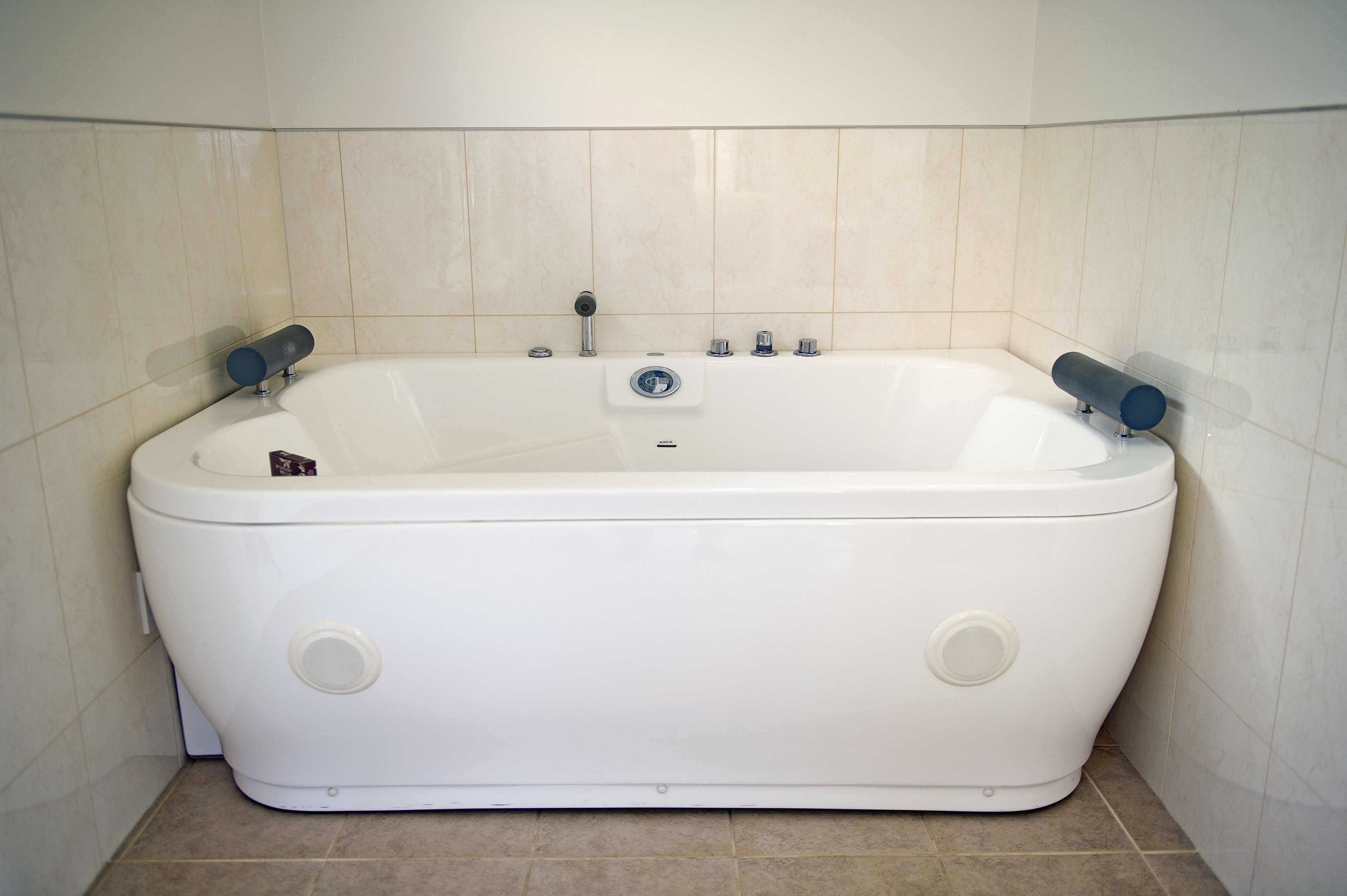 Una bañera blanca independiente con dos grifos y un panel de control, frente a paredes cubiertas de azulejos beige.