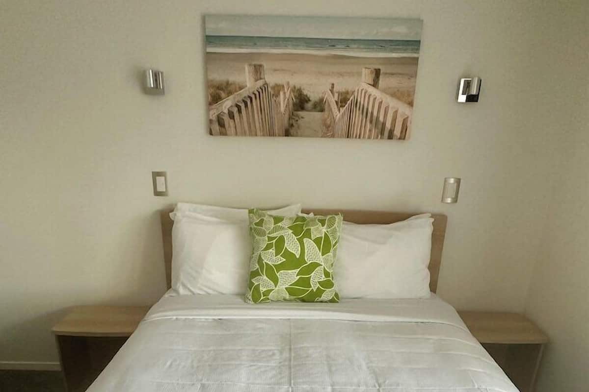 A bed with a white mattress, a green throw blanket, and a pillow with a leaf pattern. There is a painting of a beach scene on the wall above the bed.