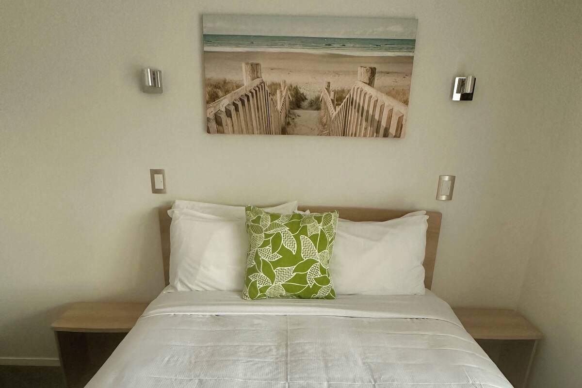 A neatly made bed with a green blanket and a pillow with a leaf pattern, a wooden headboard, and a painting of a beach scene above the bed.