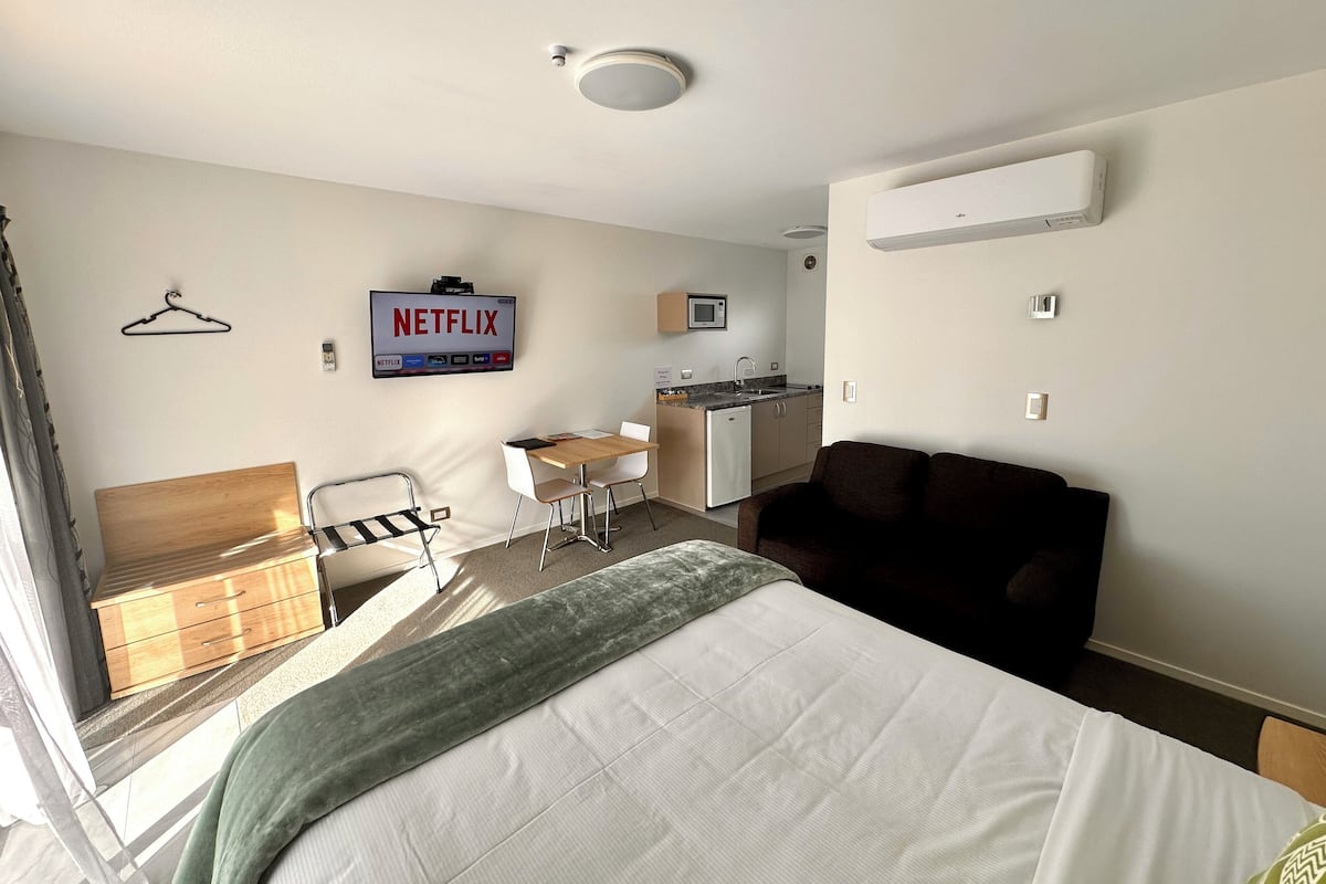 A hotel room with a bed, a desk, a chair, a small kitchenette, and a TV mounted on the wall.