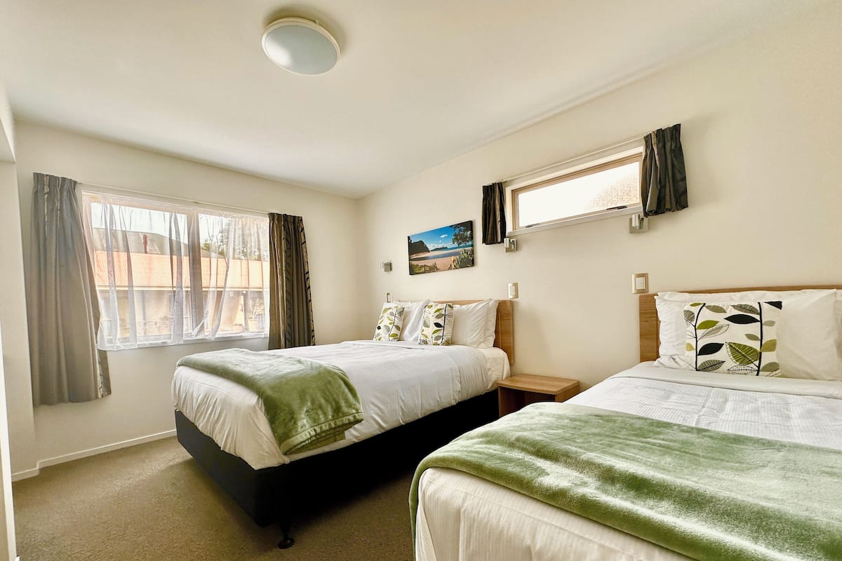 A hotel room with two beds, a window with curtains, a painting on the wall, and a ceiling light.