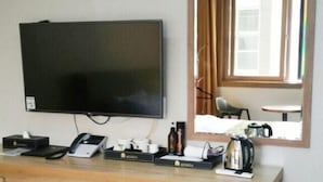 A hotel room with a flat-screen TV mounted on the wall, a wooden desk with a telephone, a box of tissues, a coffee maker, and a small table with a chair. - Benikea Central Hotel (Tongyeong)